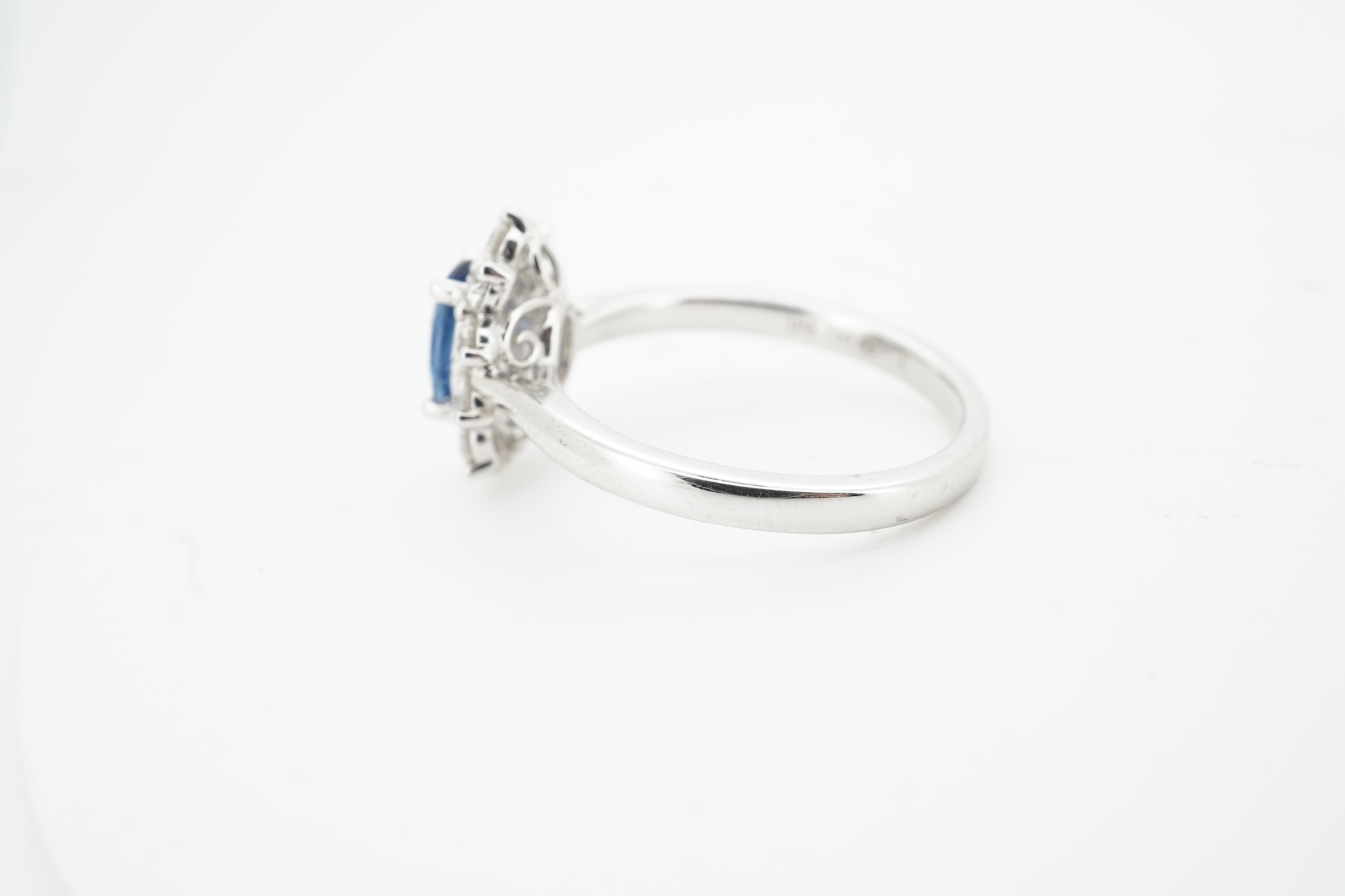 A sapphire and diamond cluster ring, claw-set with an oval sapphire weighing approximately 0.77 carats, within a border of brilliant-cut diamonds totalling approximately 0.50 carats, mounted in white gold, size M, stampe